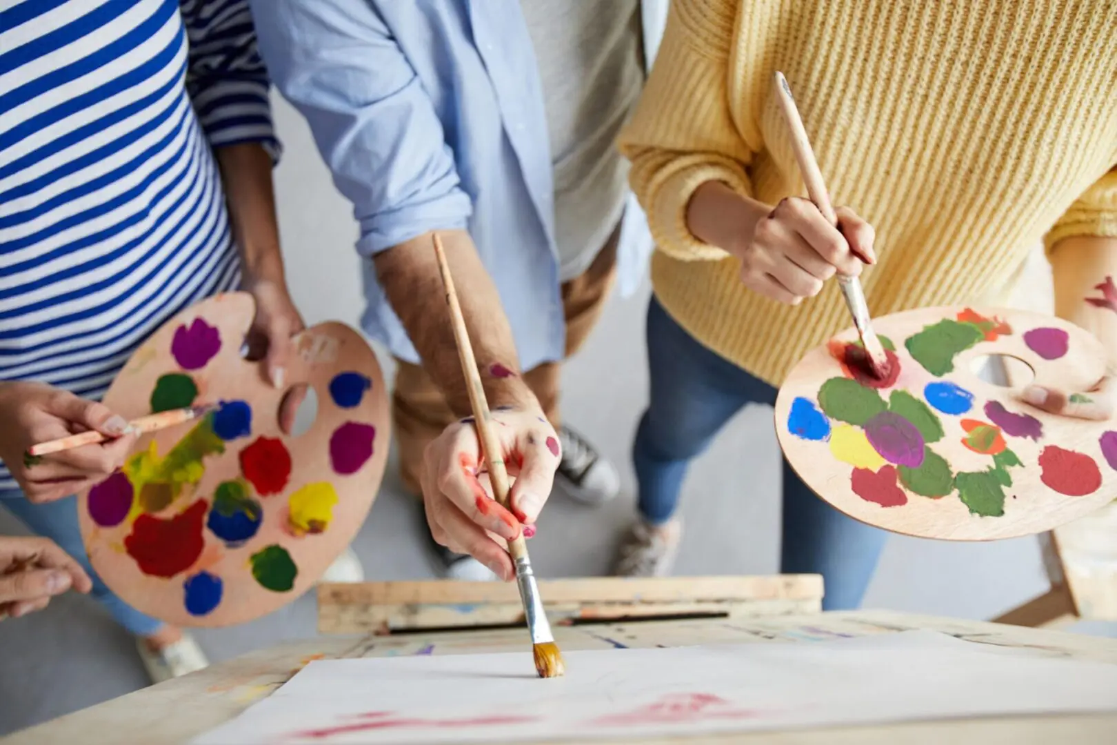 Three people hold paint palettes and brushes, standing close together, actively painting on a shared canvas.