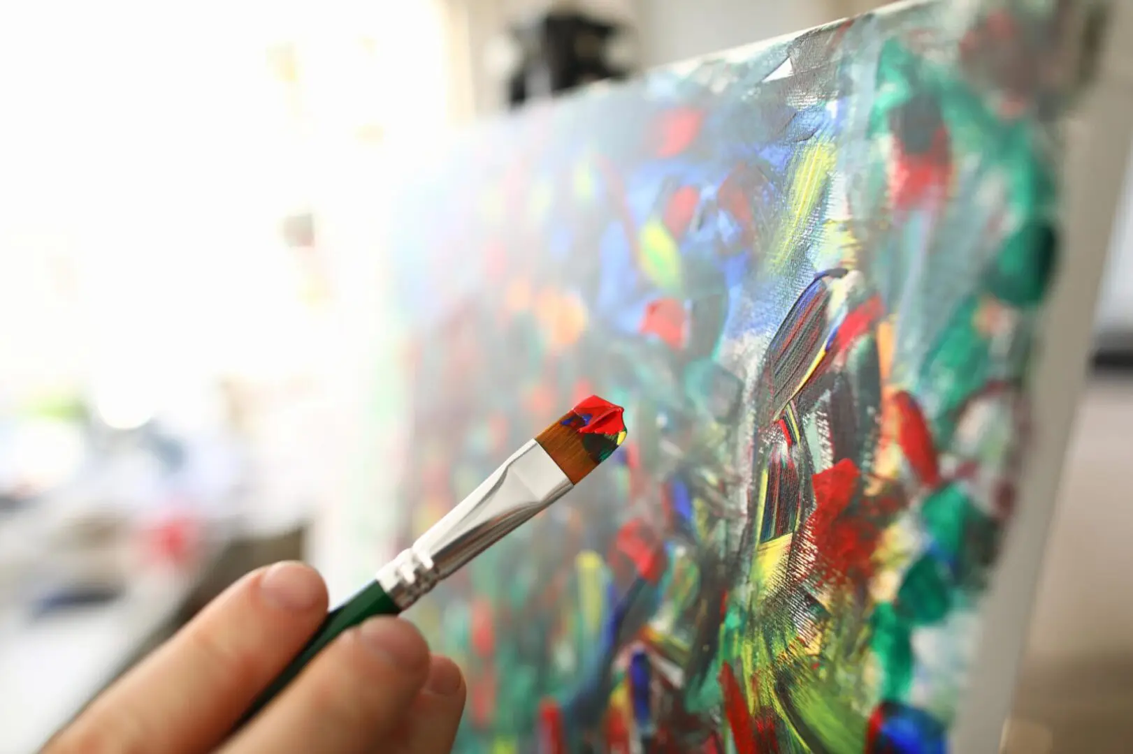 A person holds a paintbrush with red paint near a colorful abstract painting on a canvas.