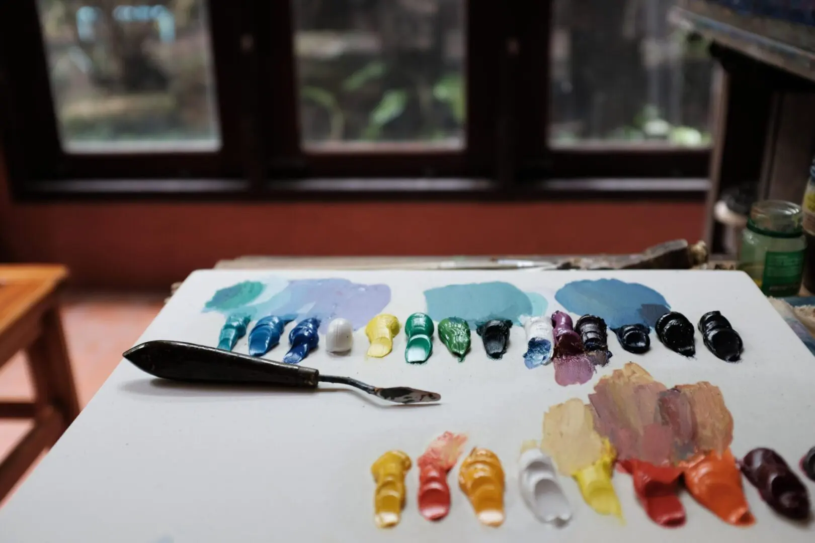 A palette knife and various paint colors arranged on a white surface in a room with large windows.