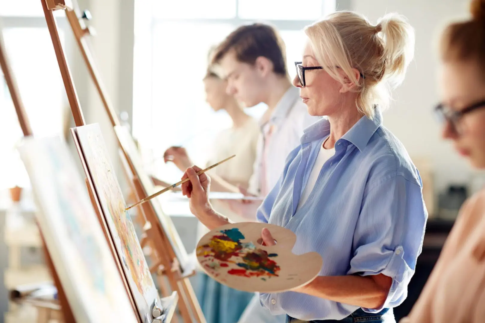 Woman painting on an easel in an art class, holding a palette. Other people are painting in the background.