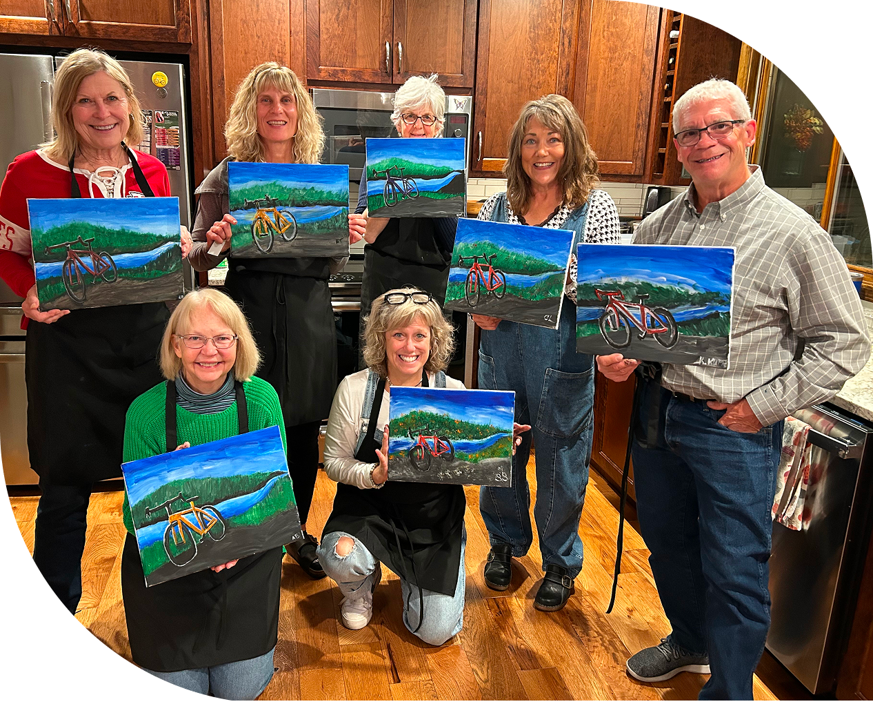 Seven people holding similar landscape paintings with a bicycle, standing in a kitchen with wooden cabinets.