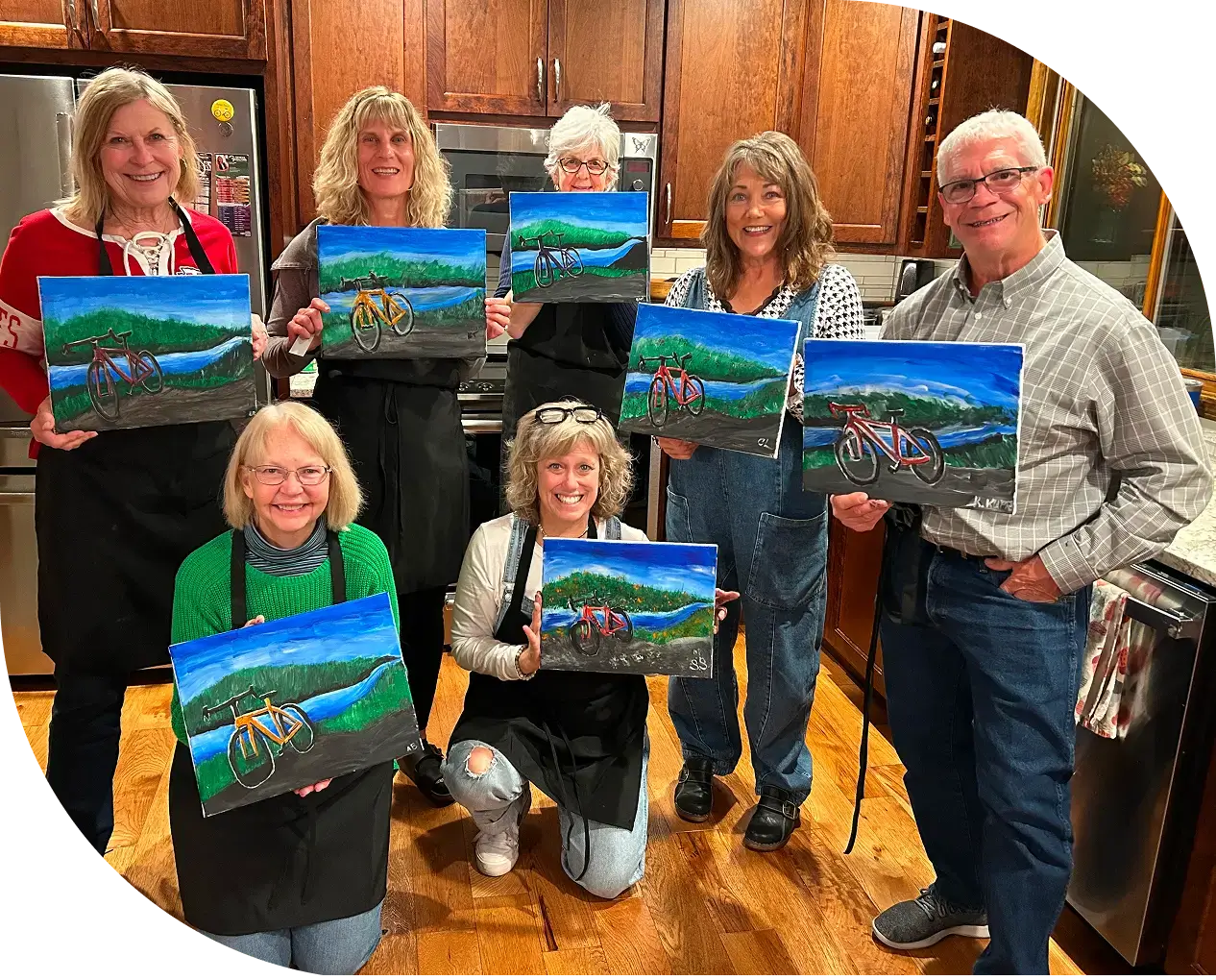 Seven people holding similar landscape paintings with a bicycle, standing in a kitchen with wooden cabinets.